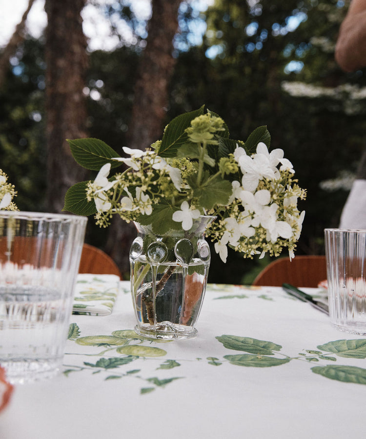 A small, handcrafted Carlotta Vase filled with white flowers sits on a table covered by a floral tablecloth. Two clear drinking glasses are nearby, with trees visible in the blurred background.