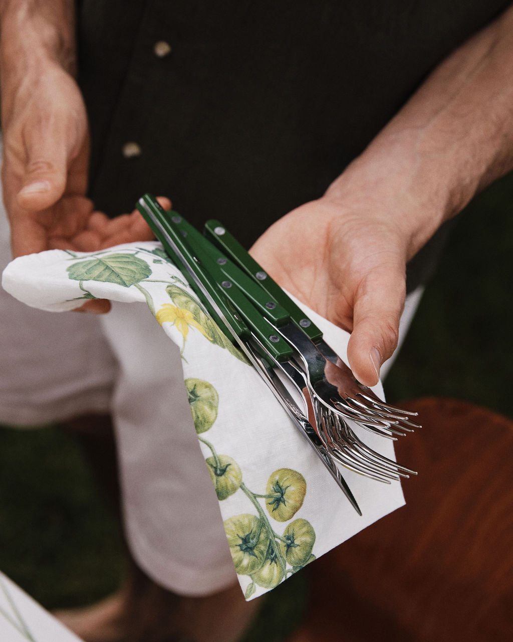 A person stands outdoors in casual attire, holding green-handled utensils wrapped in a Tomate Verte Linen Napkin from Summerill & Bishop.