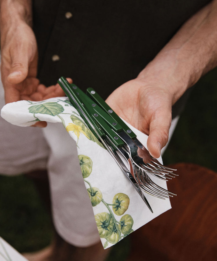 A person stands outdoors in casual attire, holding green-handled utensils wrapped in a Tomate Verte Linen Napkin from Summerill & Bishop.