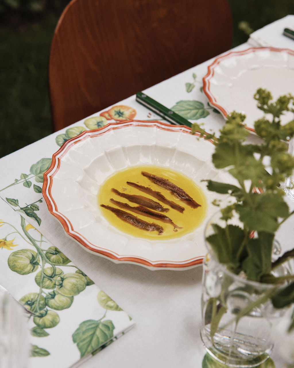 A white Eva Melamine Bowl, Italian-inspired with a scalloped rim, holds olive oil and anchovy fillets on a floral tablecloth beside a glass of green herbs. Set of 4.