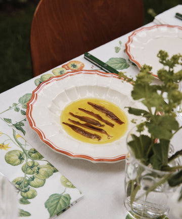 A white Eva Melamine Bowl, Italian-inspired with a scalloped rim, holds olive oil and anchovy fillets on a floral tablecloth beside a glass of green herbs. Set of 4.
