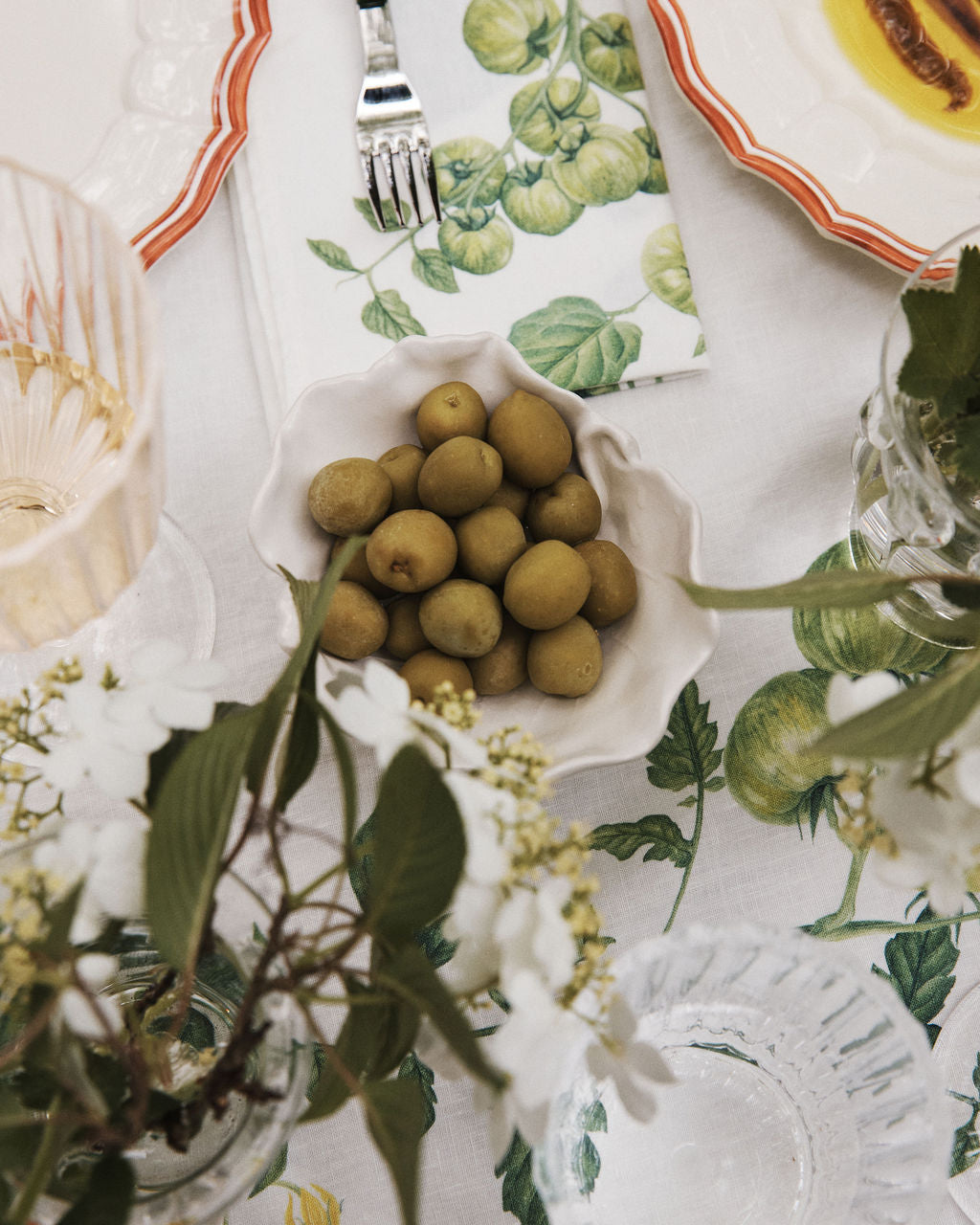 Green olives are served in a Leaf Bowl, a hand-painted earthenware piece, set on a white tablecloth among glassware, plates, and floral decorations.