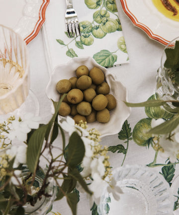 Green olives are served in a Leaf Bowl, a hand-painted earthenware piece, set on a white tablecloth among glassware, plates, and floral decorations.