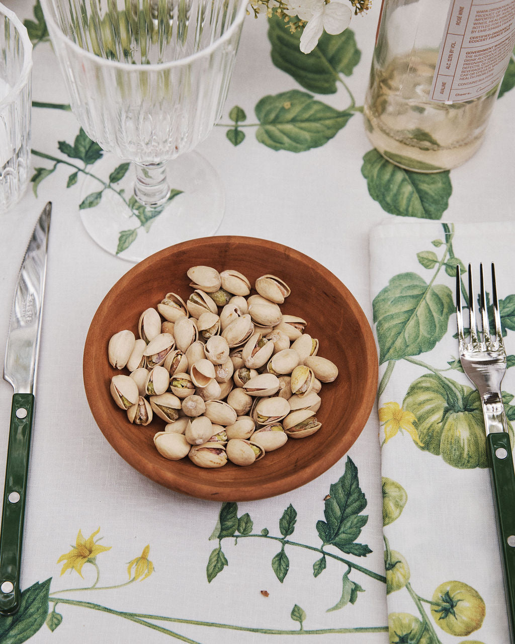 An August Wood Serving Bowl, handcrafted from American dark cherry wood and filled with pistachios, sits on a table set with a knife, fork, glassware, a bottle, and a floral-patterned tablecloth and napkin.
