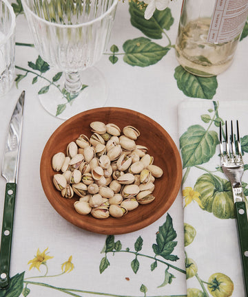 An August Wood Serving Bowl, handcrafted from American dark cherry wood and filled with pistachios, sits on a table set with a knife, fork, glassware, a bottle, and a floral-patterned tablecloth and napkin.