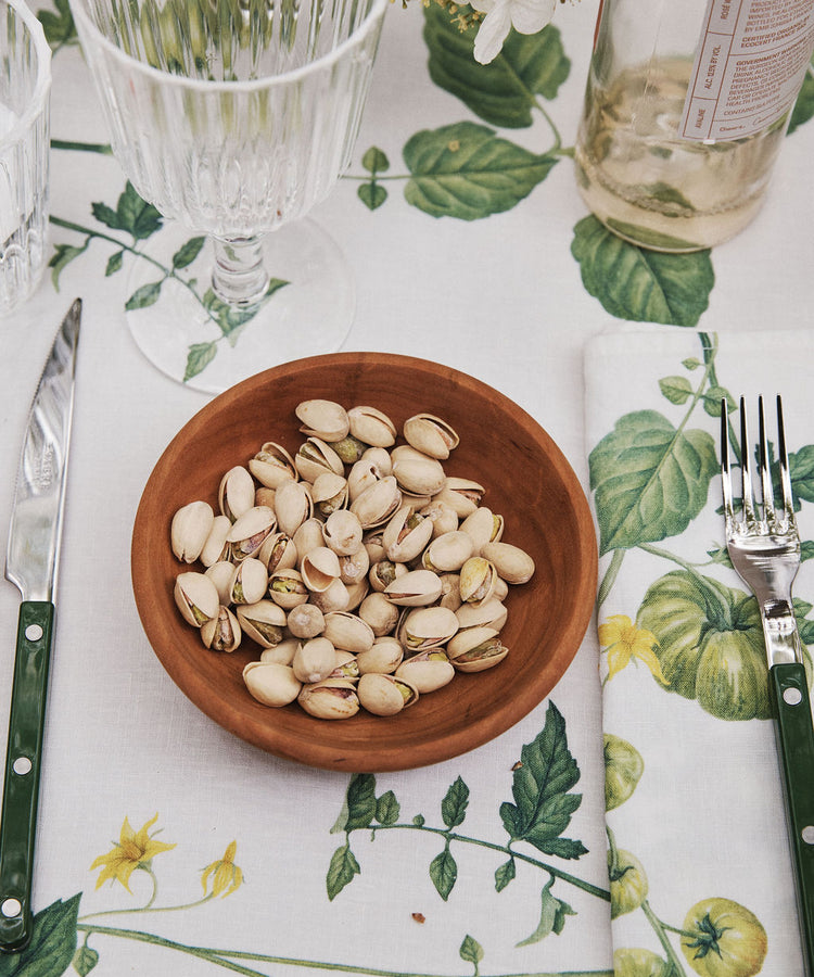 An August Wood Serving Bowl, handcrafted from American dark cherry wood and filled with pistachios, sits on a table set with a knife, fork, glassware, a bottle, and a floral-patterned tablecloth and napkin.