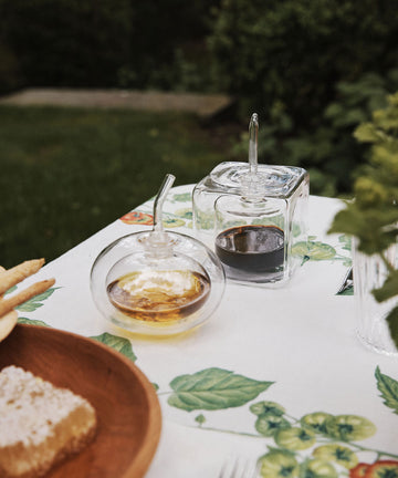 The Cube Oil & Vinegar Cruet—comprised of a round, double-walled vessel filled with oil and a square one holding vinegar—rests atop a patterned tablecloth beside a wooden plate of bread, outdoors, evoking an inviting kitchen atmosphere.