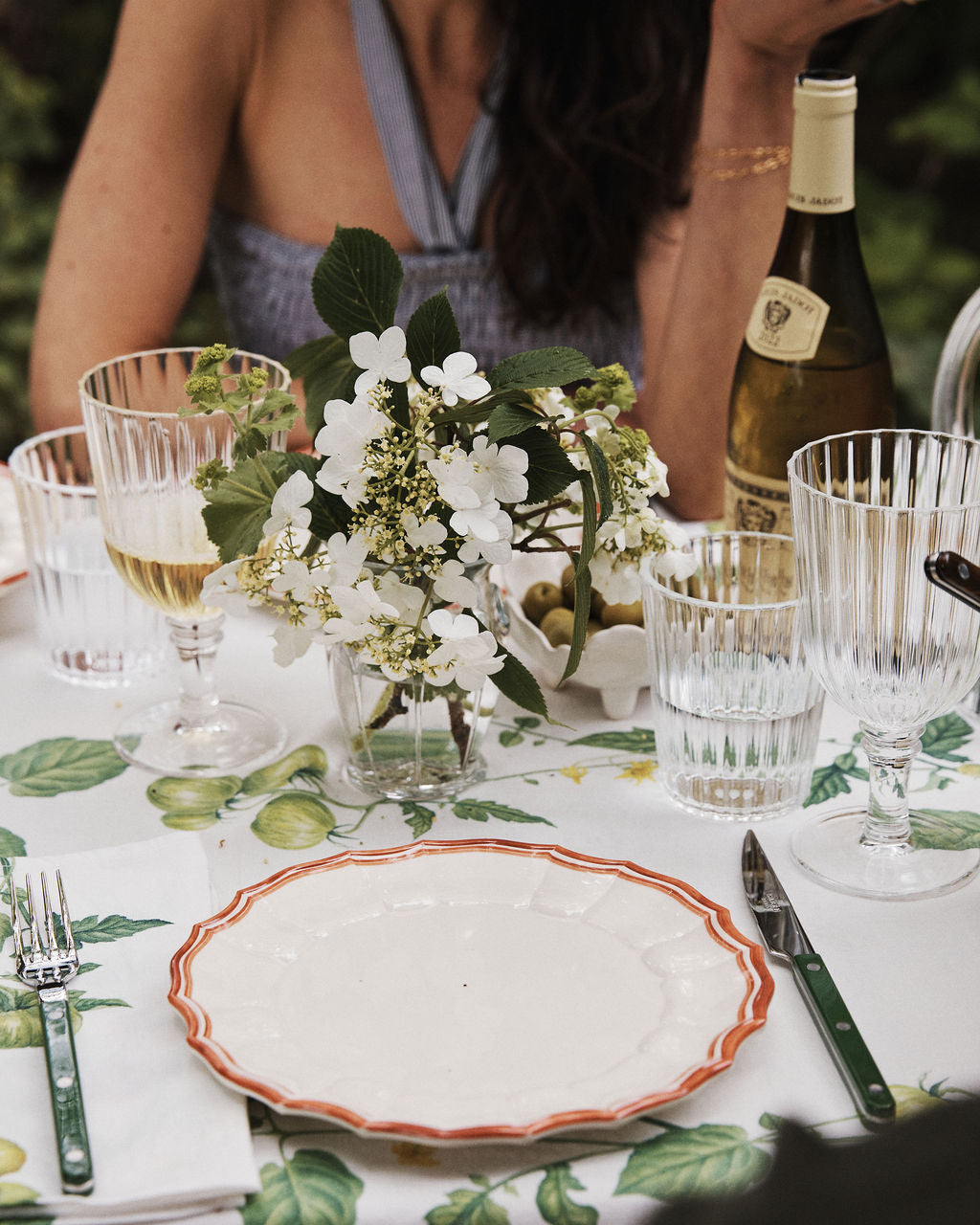 A meal table set with Eva Melamine Small Plate, Set of 4—BPA-free plates with a scalloped red trim—plus cutlery, glasses, wine, and white flowers in a vase; a person is seated in the background.
