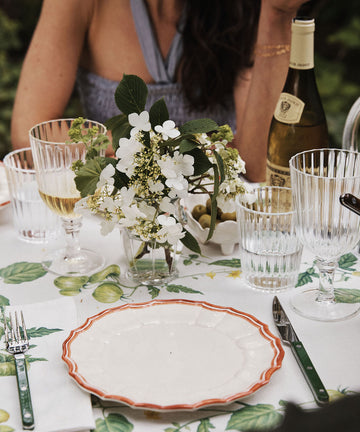 A meal table set with Eva Melamine Small Plate, Set of 4—BPA-free plates with a scalloped red trim—plus cutlery, glasses, wine, and white flowers in a vase; a person is seated in the background.