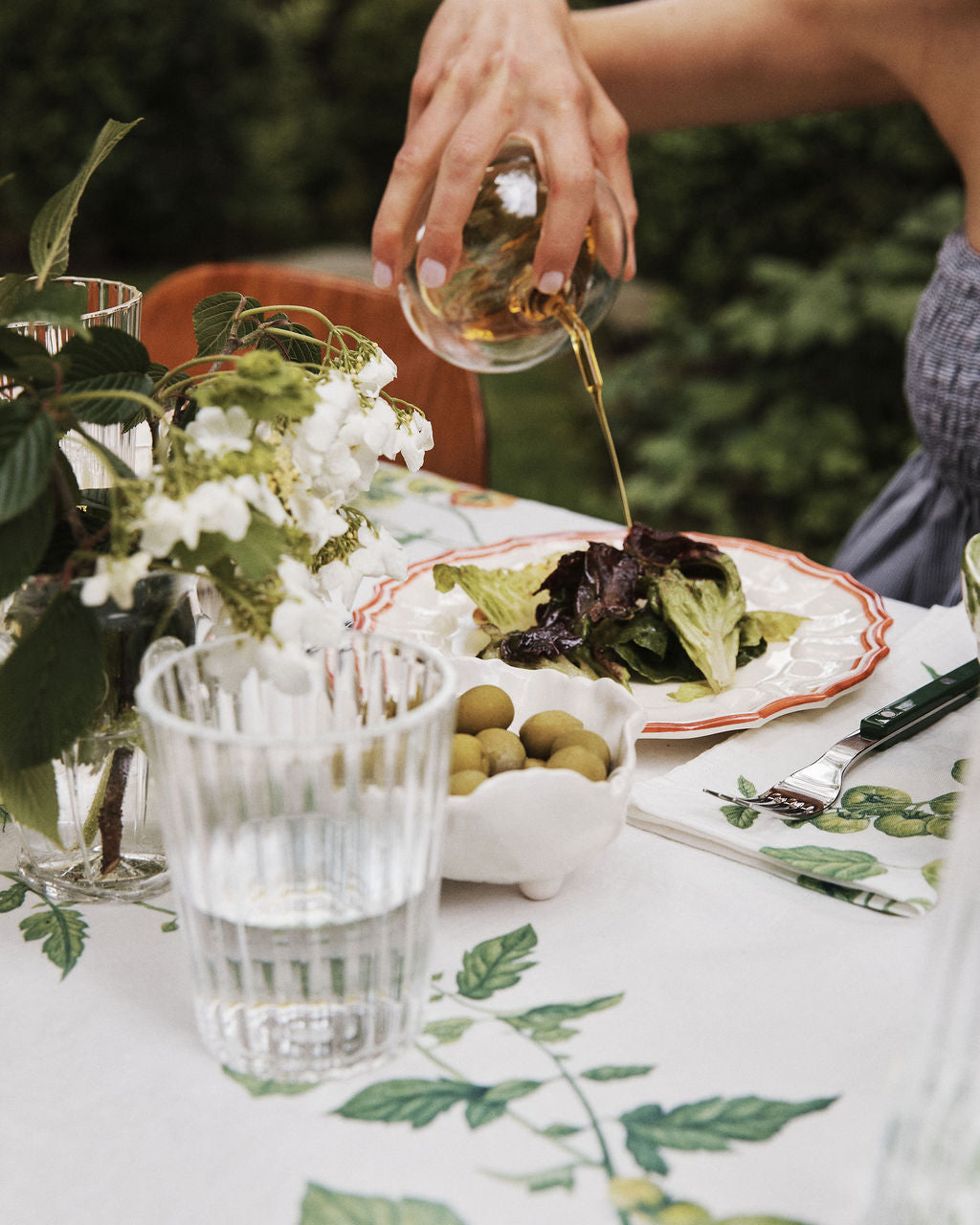 Using the Cylinder Oil & Vinegar Cruet, a person pours dressing onto a plate of salad at an outdoor table set with a glass of water, a bowl of green olives, and a vase of white flowers.