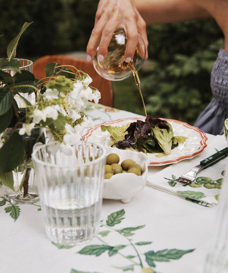 Using the Cylinder Oil & Vinegar Cruet, a person pours dressing onto a plate of salad at an outdoor table set with a glass of water, a bowl of green olives, and a vase of white flowers.