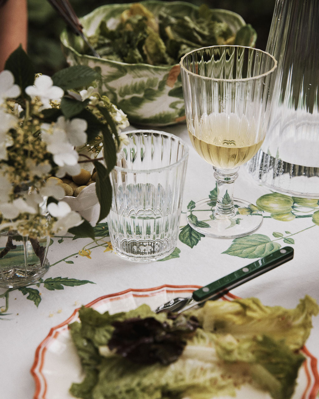 Table set with a plate of mixed greens, a fork, white wine, a Fresco Tumbler from the Set of 4 for water, floral arrangement, and a bowl of salad on a lemon-patterned tablecloth.