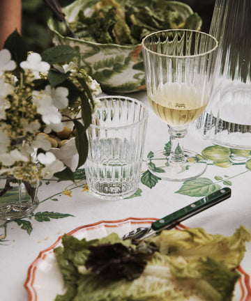 Table set with a plate of mixed greens, a fork, white wine, a Fresco Tumbler from the Set of 4 for water, floral arrangement, and a bowl of salad on a lemon-patterned tablecloth.