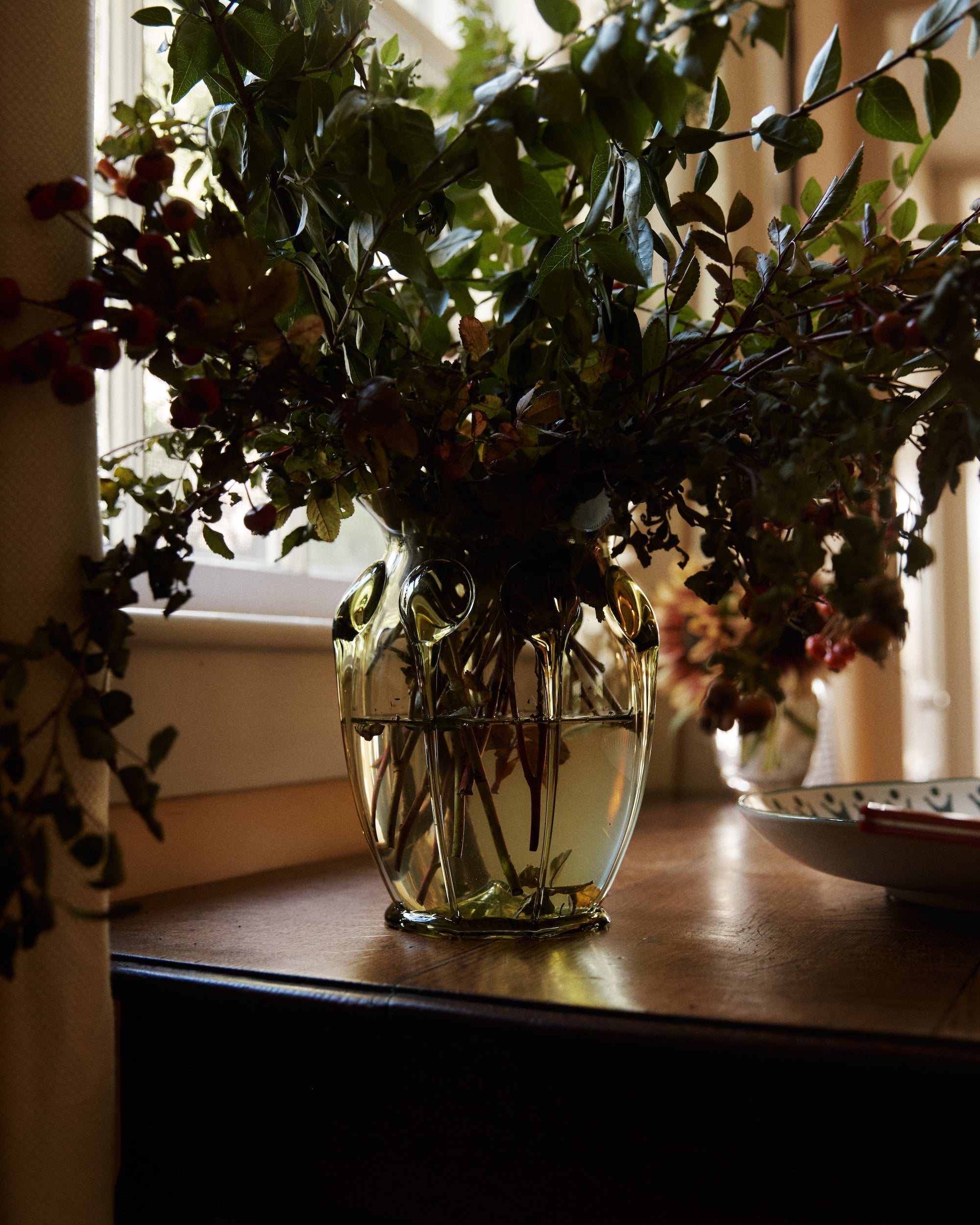 A Carlotta Vase filled with water and green branches sits on a wooden table by a window in natural light.