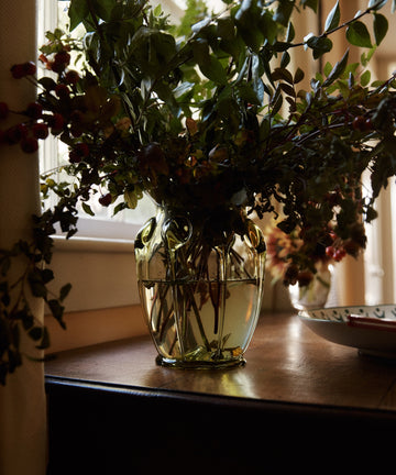 A Carlotta Vase filled with water and green branches sits on a wooden table by a window in natural light.