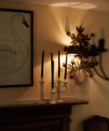 A mantel with Old World charm displays the Loretta Candelabra holding black candles, a large swan drawing, and a leafy wall sconce with a shell accent above.