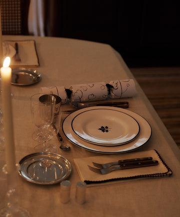 A formal dining table with a candle, glassware, cutlery, and the Beatrix Dinner Plate, Set of 4 featuring a blue-and-white botanical design on a cream tablecloth.