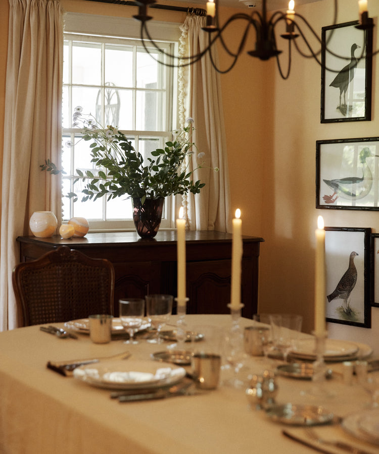 A dark plum Aura Vase holding green stems and white flowers, placed on a wooden sideboard behind a candlelit dining table.