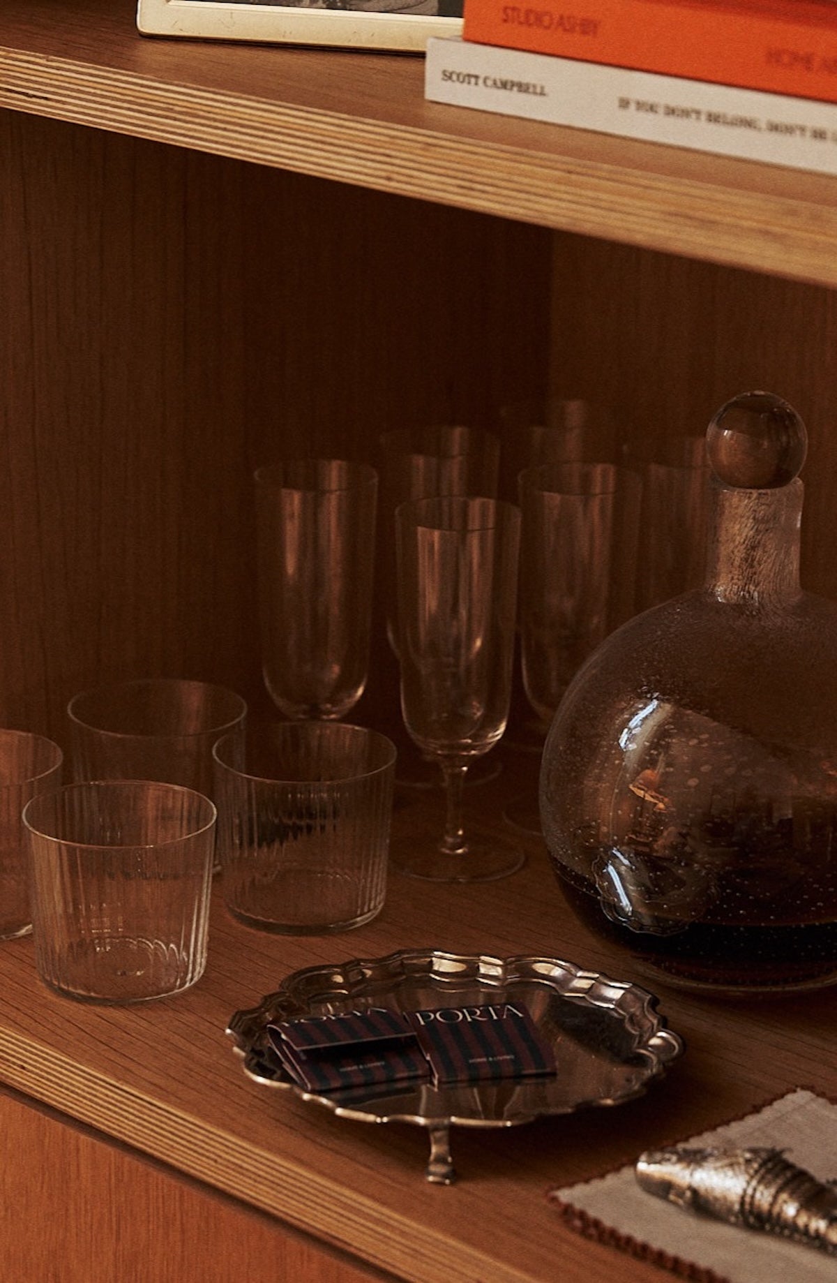 A wooden shelf showcases the Venezia Champagne Flute, Set of 6—timeless drinkware beside a glass decanter with dark liquid, a silver tray holding matchboxes, and part of a white cloth.