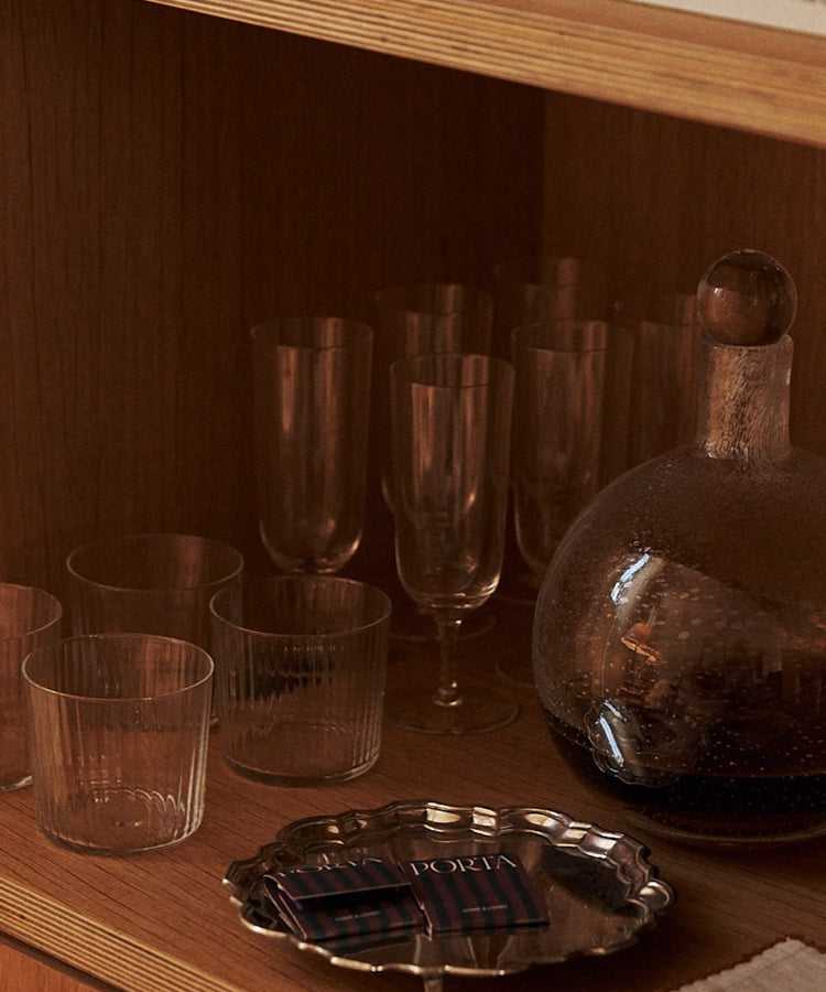 A wooden shelf showcases the Venezia Champagne Flute, Set of 6—timeless drinkware beside a glass decanter with dark liquid, a silver tray holding matchboxes, and part of a white cloth.