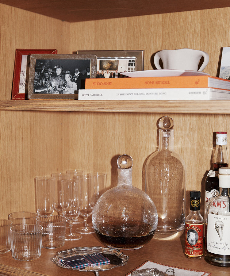 Wooden shelf with framed photos, books, glassware, a Bubble Glass Carafe, two glass decanters, liquor bottles, a silver tray, and a coaster featuring a black and white image.