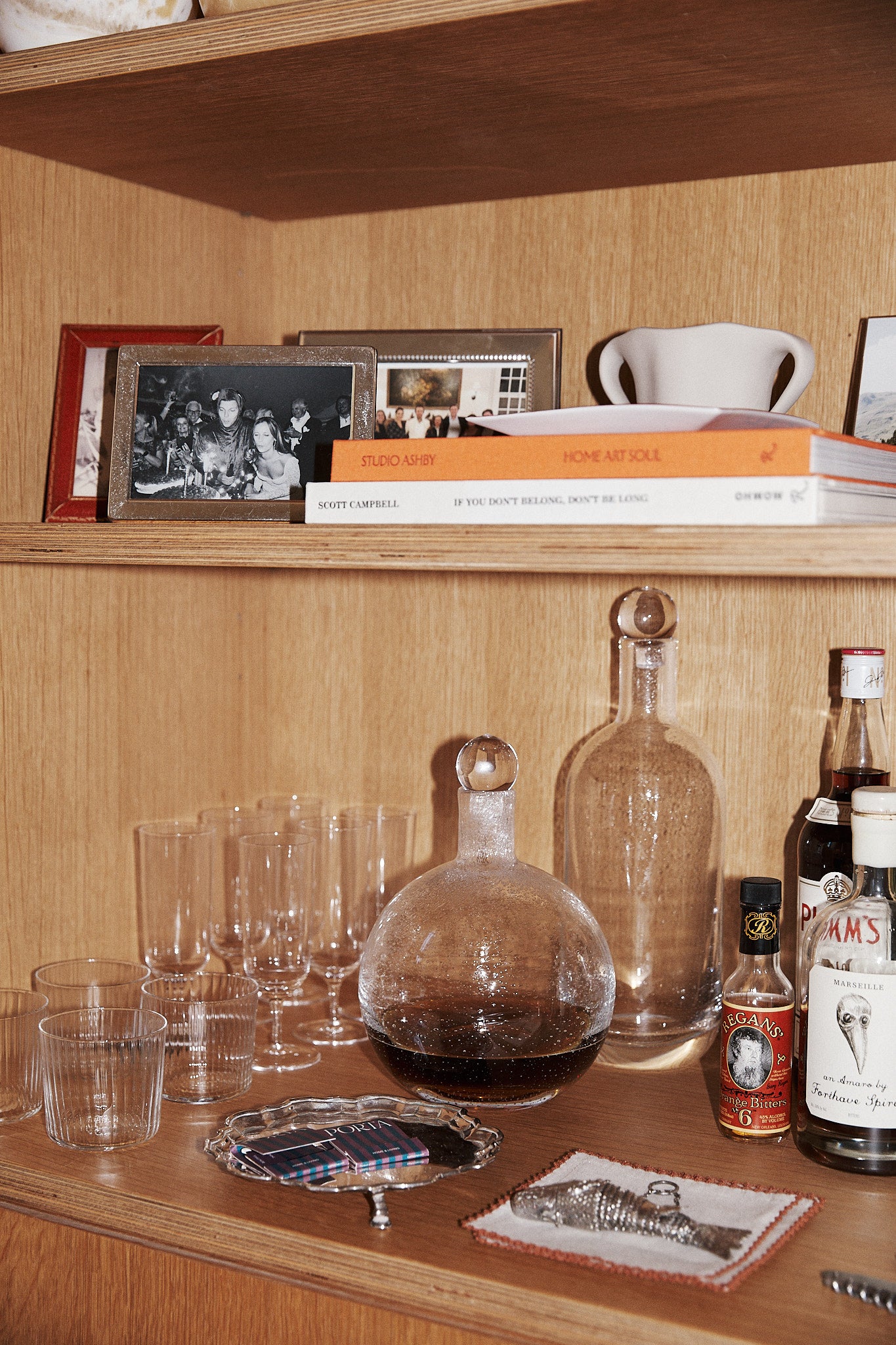 Wooden shelf featuring elegant glassware, a hand-blown Bubble Glass Carafe, two decanters, assorted liquor bottles, framed photos, books, a silver tray, and a vintage photo coaster.