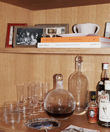 Wooden shelf featuring elegant glassware, a hand-blown Bubble Glass Carafe, two decanters, assorted liquor bottles, framed photos, books, a silver tray, and a vintage photo coaster.