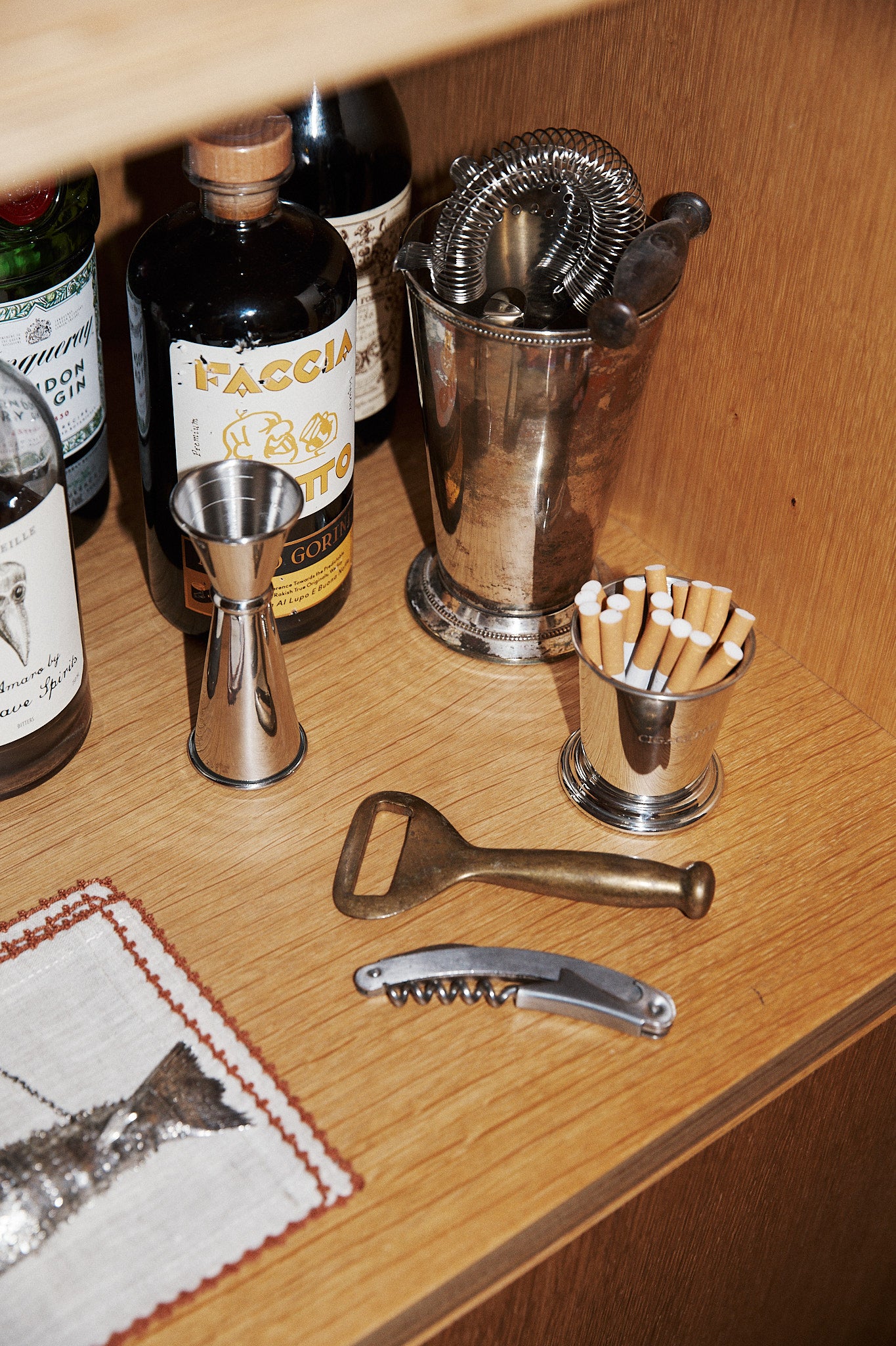 The Cleo Cigarette Cup, liquor bottles, various bar tools, and a cocktail strainer are arranged on a wooden shelf.