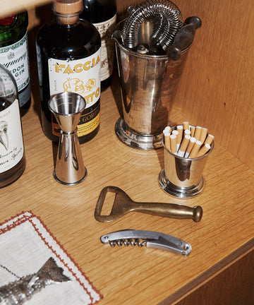 The Cleo Cigarette Cup, liquor bottles, various bar tools, and a cocktail strainer are arranged on a wooden shelf.