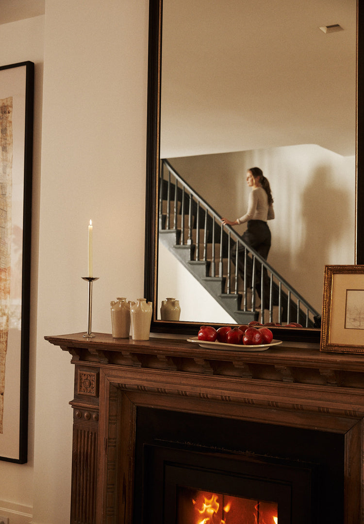 Woman standing on a staircase reflected in a mirror above a fireplace.