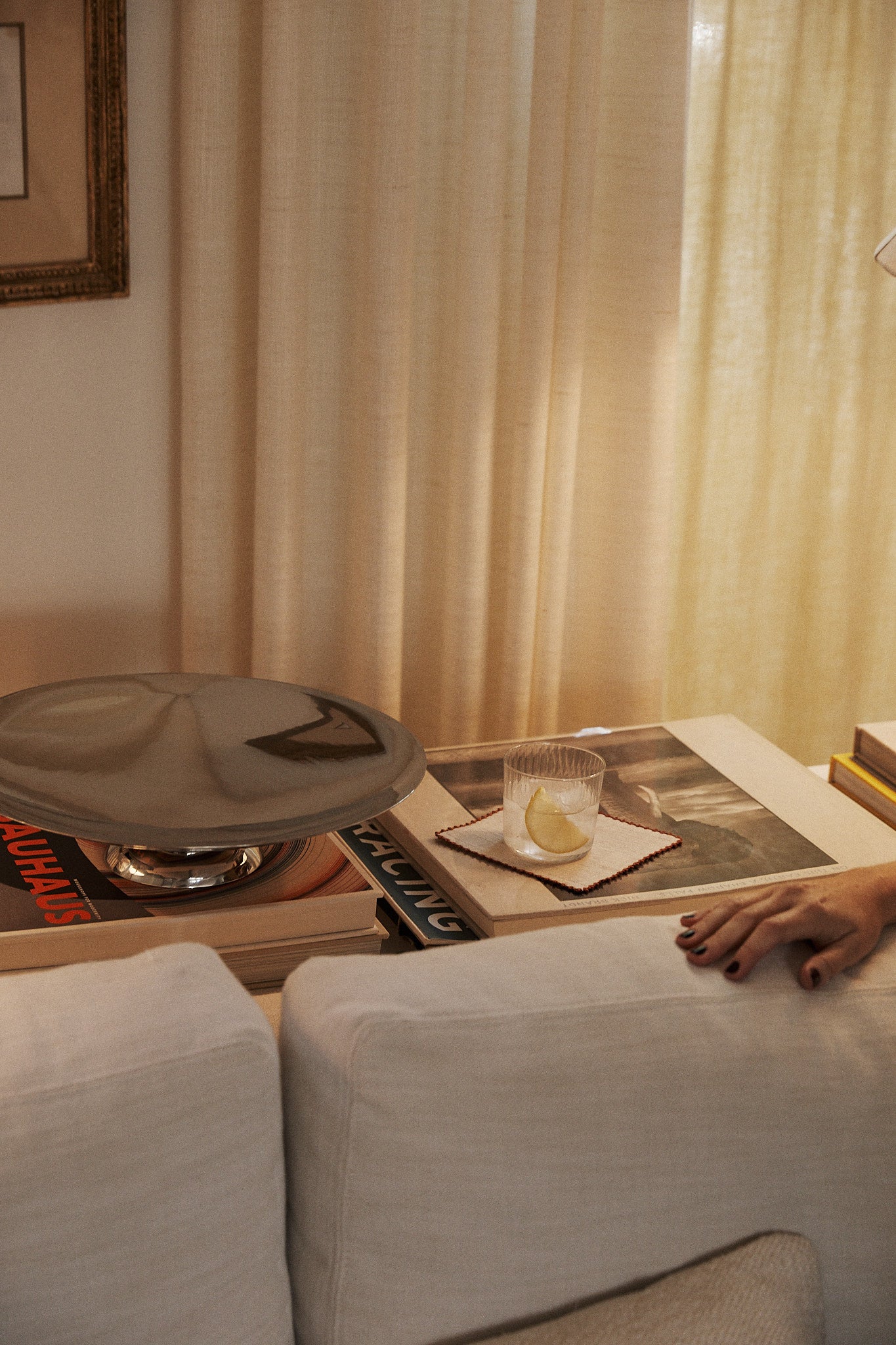 A glass of water with lemon rests on coasters atop books behind a white sofa, near a person's hand and a set of Nico Cocktail Napkins, Set of 4, featuring hand-finished details and subtle contrast stitching.