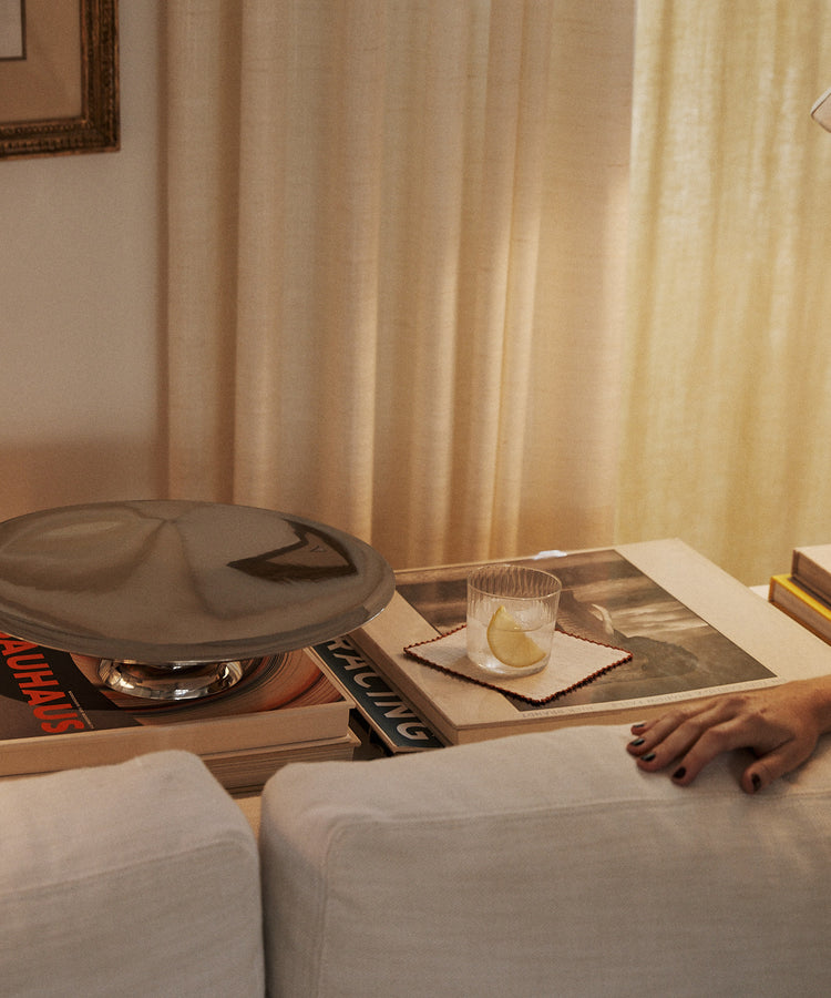 A glass of water with lemon rests on coasters atop books behind a white sofa, near a person's hand and a set of Nico Cocktail Napkins, Set of 4, featuring hand-finished details and subtle contrast stitching.