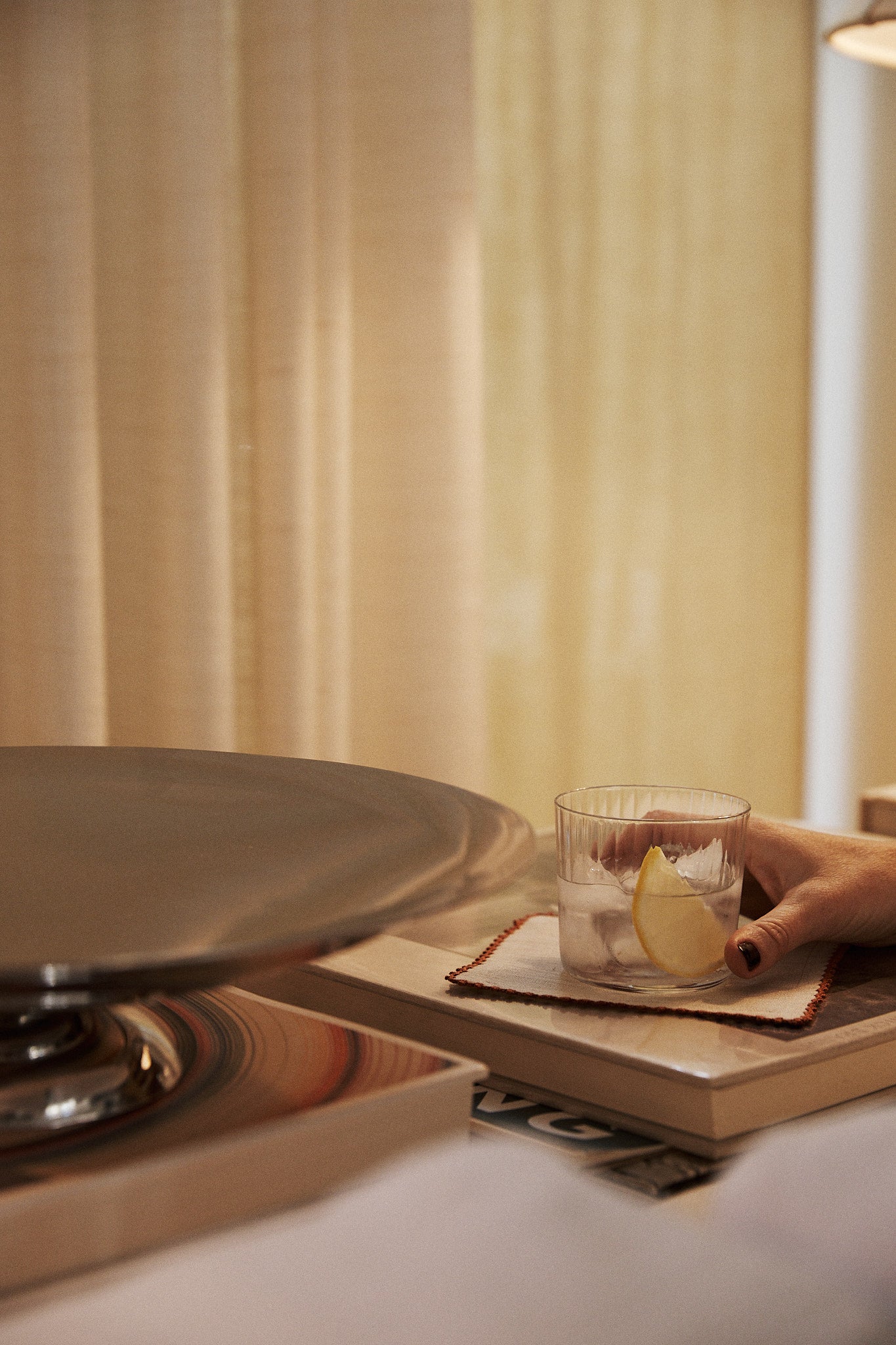 A hand holds a Teo Ridged Tumbler filled with iced water and a lemon slice, the glass resting on a coaster atop a closed book near a metal tray and beige curtains.