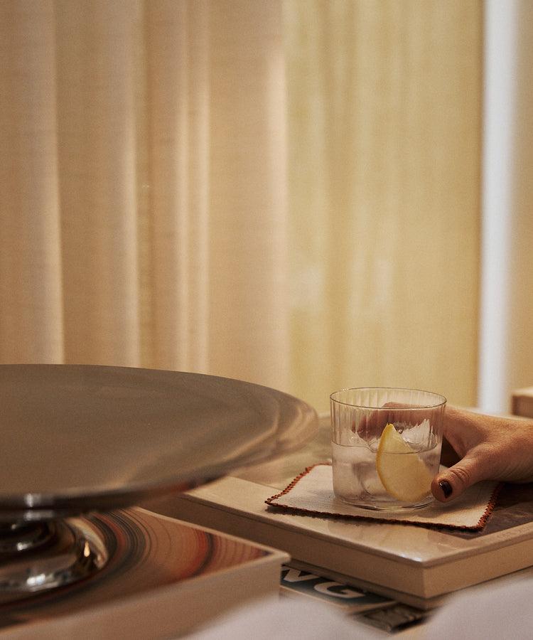 A hand holds a Teo Ridged Tumbler filled with iced water and a lemon slice, the glass resting on a coaster atop a closed book near a metal tray and beige curtains.