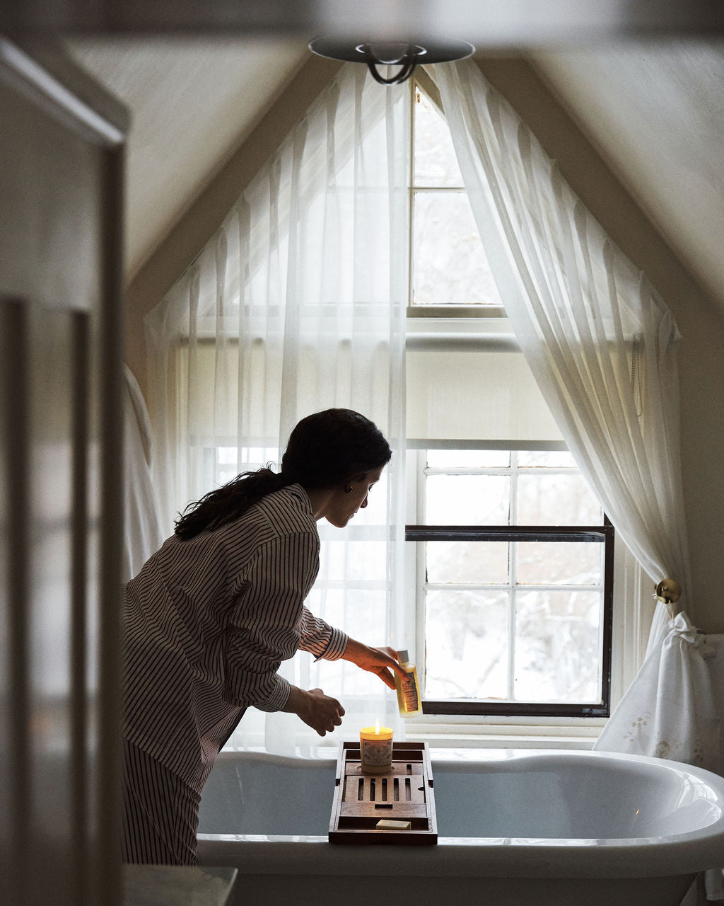 A person in striped pajamas places Dermoprotective Oil and a candle on a wooden bath tray over a white tub, preparing for soothing hydration and delicate skin care by the window with sheer curtains.