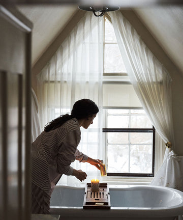 A person in striped pajamas places Dermoprotective Oil and a candle on a wooden bath tray over a white tub, preparing for soothing hydration and delicate skin care by the window with sheer curtains.
