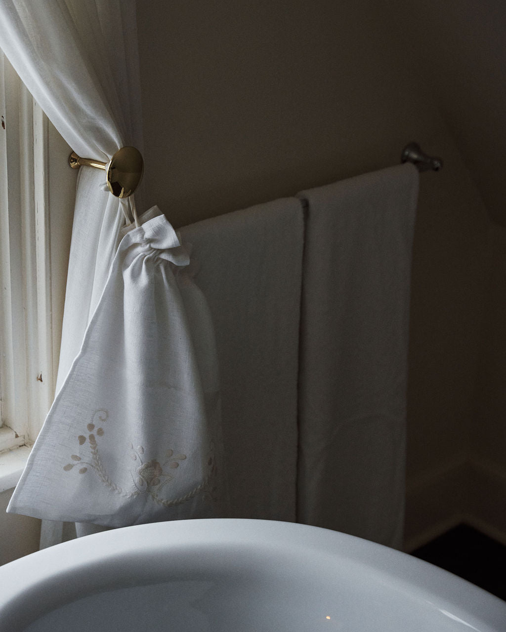 A linen drawstring bag beside a window, paired with white towels by a white sink in a softly lit bathroom, captures the handmade charm and elegant essence of Viana do Castelo embroidery.