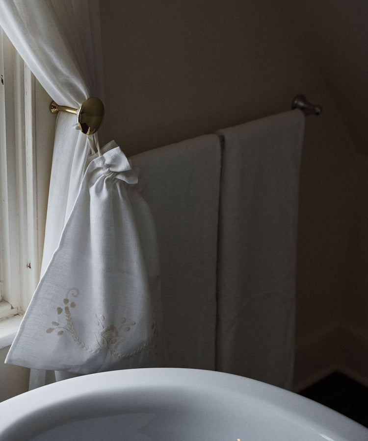 A linen drawstring bag beside a window, paired with white towels by a white sink in a softly lit bathroom, captures the handmade charm and elegant essence of Viana do Castelo embroidery.