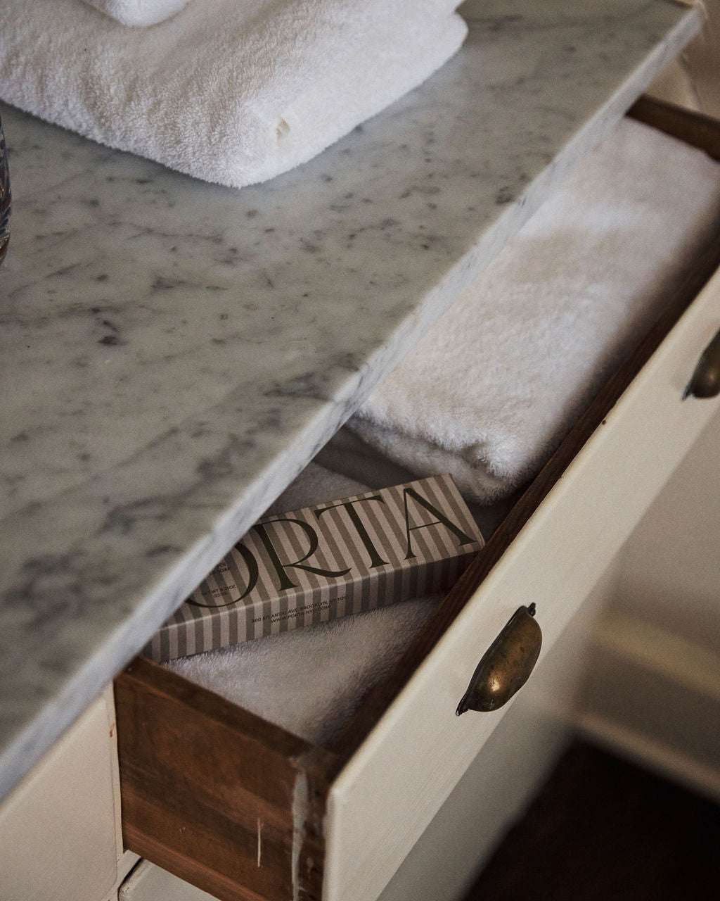 A marble countertop holds folded white towels; below, a drawer reveals more towels and a rectangular striped Porta Soap Box Gift Set, ideal for gifting wildflower-scented soaps.