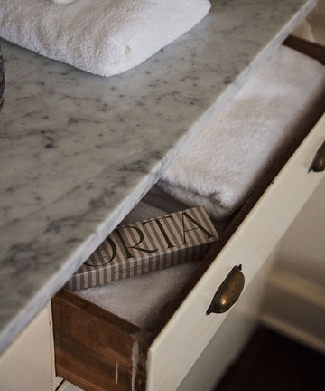 A marble countertop holds folded white towels; below, a drawer reveals more towels and a rectangular striped Porta Soap Box Gift Set, ideal for gifting wildflower-scented soaps.