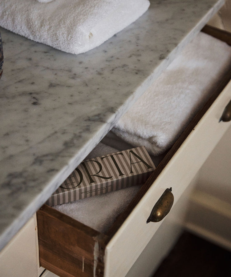 A marble countertop holds folded white towels; below, a drawer reveals more towels and a rectangular striped Porta Soap Box Gift Set, ideal for gifting wildflower-scented soaps.