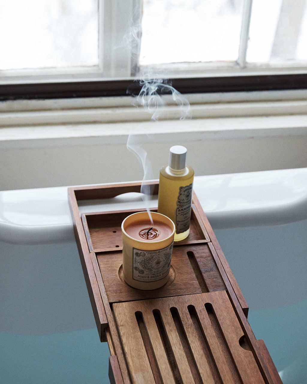 A lit Acqua Della Regina Candle sits on a wooden bath tray over a white tub by the window, filling the room with its delicate Florentine fragrance.