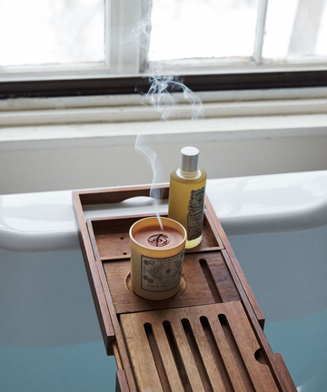 A lit Acqua Della Regina Candle sits on a wooden bath tray over a white tub by the window, filling the room with its delicate Florentine fragrance.