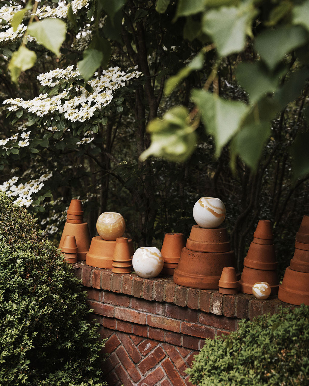 The Akila Candleholder, inspired by Egyptian Alabaster, along with terracotta pots and marbled spheres, is displayed on a brick garden wall amid green foliage and blooming branches.