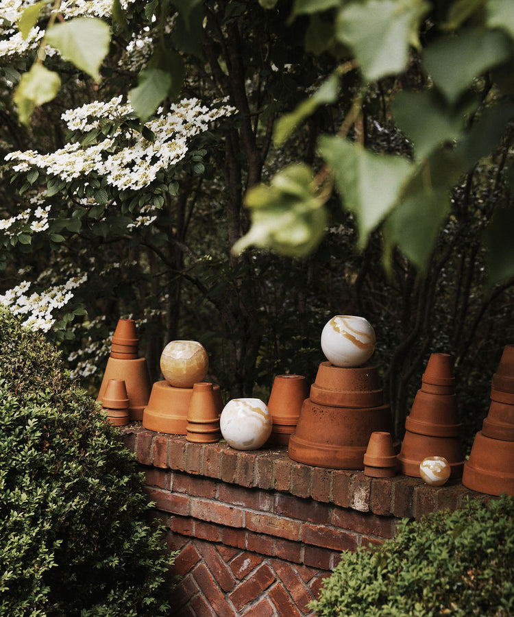 The Akila Candleholder, inspired by Egyptian Alabaster, along with terracotta pots and marbled spheres, is displayed on a brick garden wall amid green foliage and blooming branches.