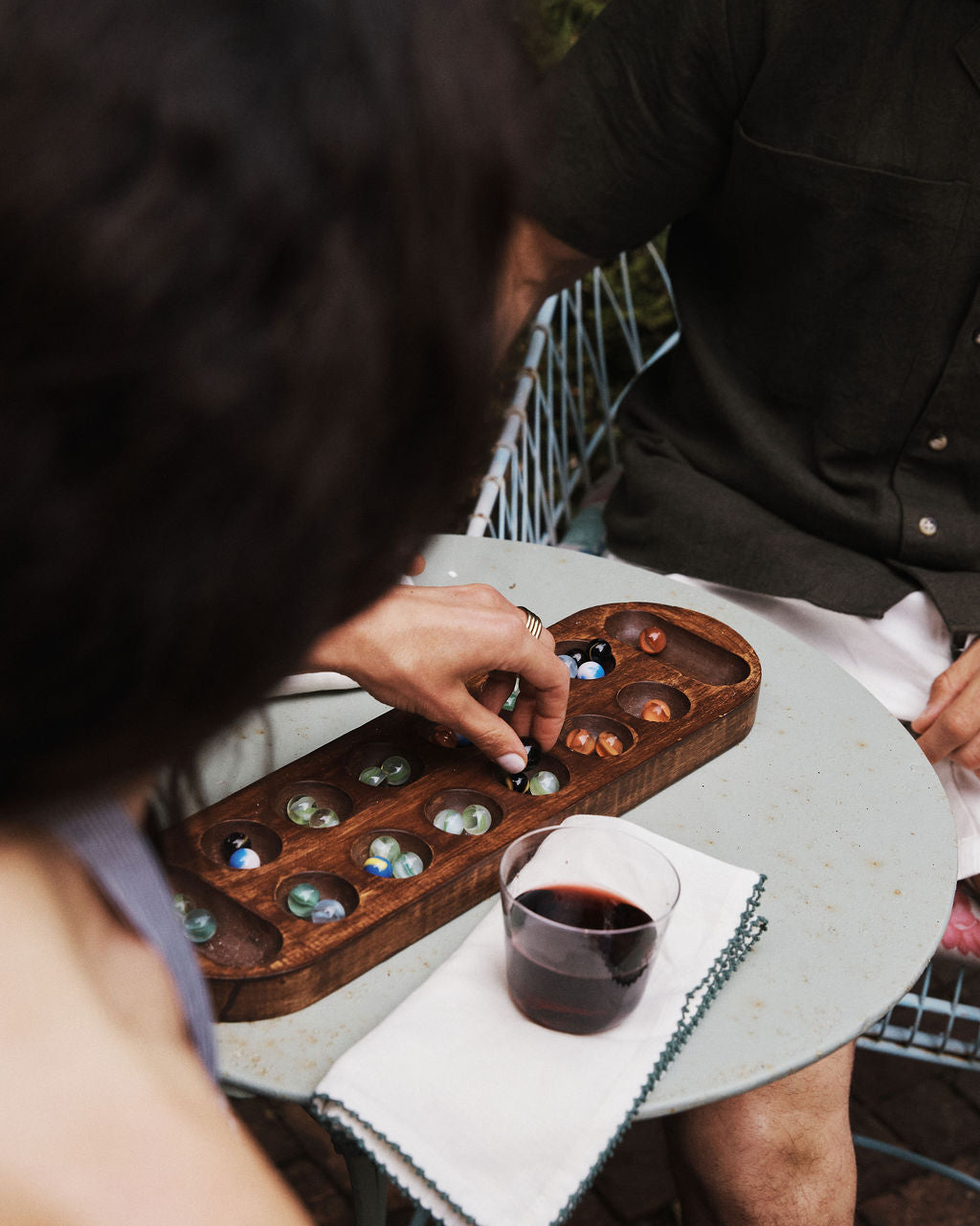 Two people sit at a round table playing mancala. One hand moves a marble on the board. A glass of red wine, an elegant table setting featuring olive oil, and a neatly folded Nico Napkin from the Set of 6 are on the table.