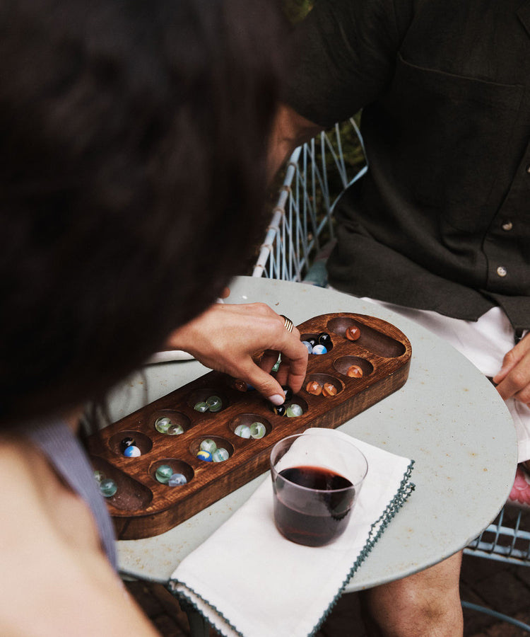 Two people sit at a round table playing mancala. One hand moves a marble on the board. A glass of red wine, an elegant table setting featuring olive oil, and a neatly folded Nico Napkin from the Set of 6 are on the table.