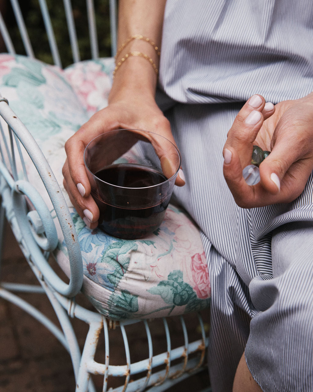 A person sits on a floral-cushioned chair, holding a plastic cup of red wine and wishing for everyday glassware like the Arc Tumbler, Set of 4—stackable tumblers made from lead-free glass.