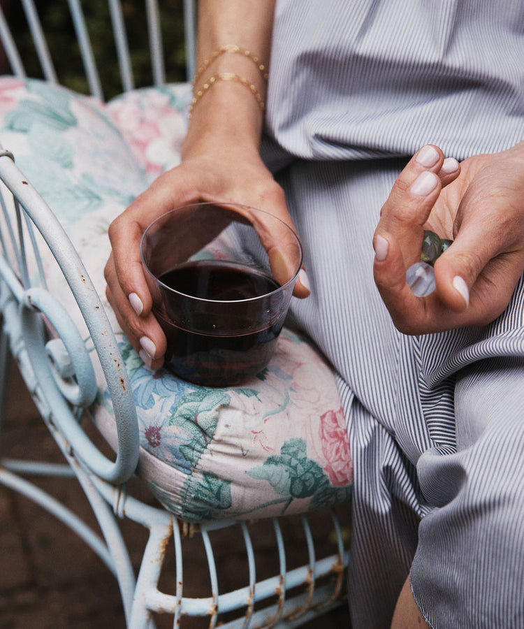 A person sits on a floral-cushioned chair, holding a plastic cup of red wine and wishing for everyday glassware like the Arc Tumbler, Set of 4—stackable tumblers made from lead-free glass.