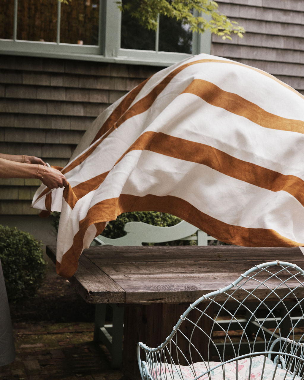 A person spreads the Riga Tablecloth, featuring hand-printed wide brown stripes, over a wooden outdoor table next to a metal chair and house siding.